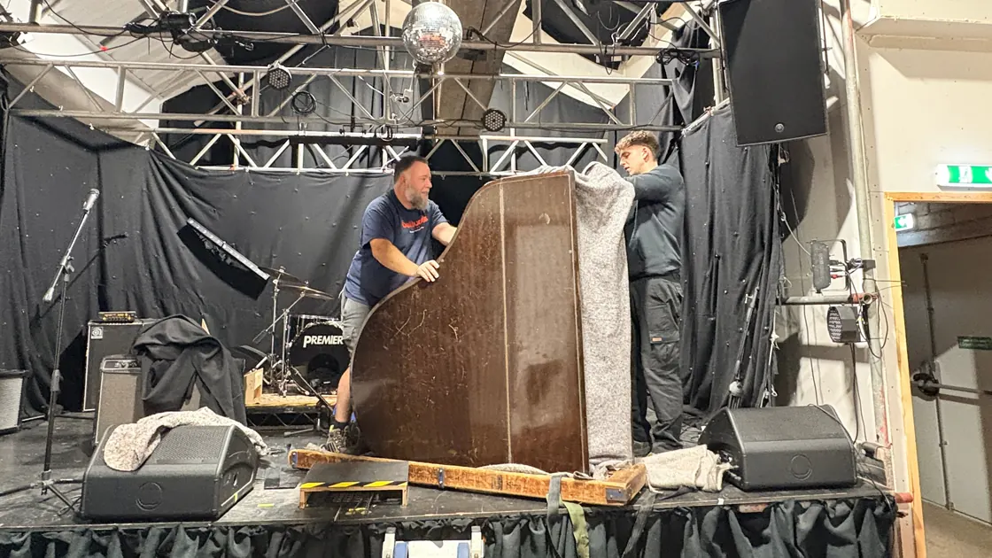 Two Yorkshire Piano Logistics movers sheeting a small grand piano on its side in its skid on the stage at Mary St Live, Sheffield, between stage monitors and a drum kit