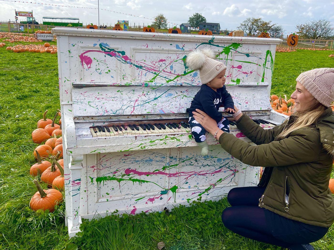 An upright piano painted white and splattered with colour, standing in a field of pumpkins with a small child sat on the keys