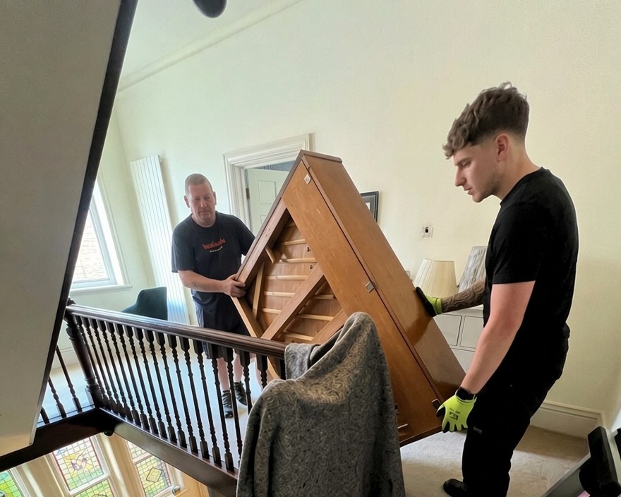 Two movers carrying an upright piano down a staircase inside a house in York