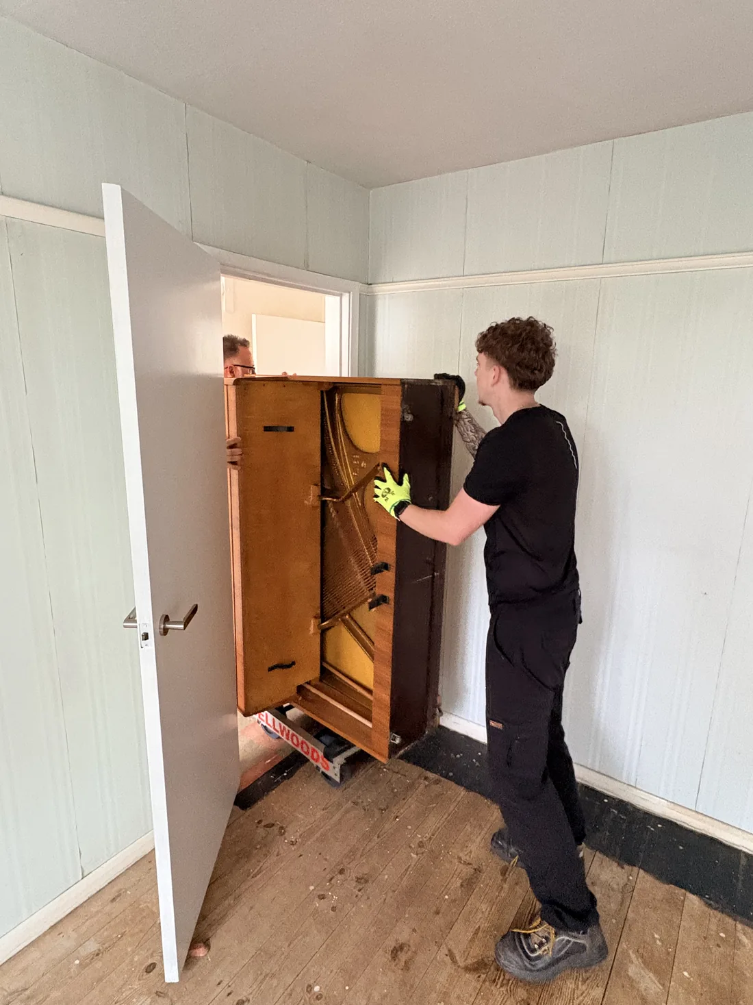 Upright piano being manoeuvred out through a narrow doorway on a piano skid