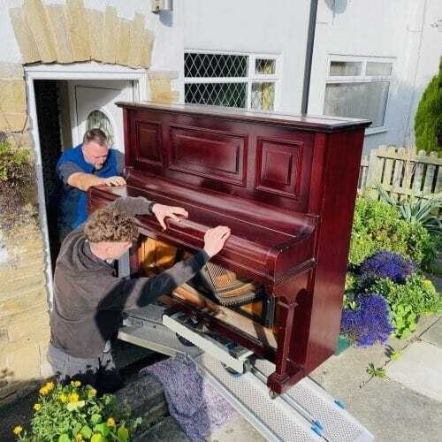 Two movers carefully bringing an upright piano out of a Yorkshire stone cottage